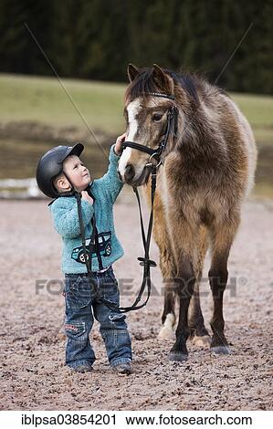 "Young child wearing a riding helmet stroking a pony, dun, with a bridle, Tyrol, Austria, Europe" View Large Photo Image Stock Image - "Young child wearing a riding helmet stroking a pony, dun, with a bridle, Tyrol, Austria, Europe". Fotosearch