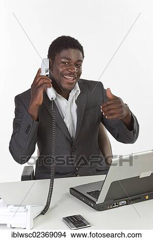 Picture - "Young dark-skinned man sitting at a laptop and holding a phone, smiling while making a thumbs-up gesture". Fotosearch
