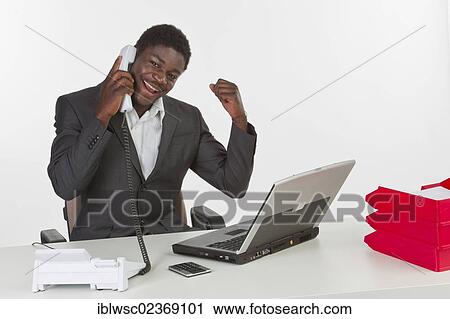 Young dark-skinned man sitting at a laptop and holding a phone while looking happy View Large Photo Image Stock Image - Young dark-skinned man sitting at a laptop and holding a phone while looking happy. Fotosearch