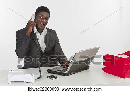 "Young dark-skinned man sitting at a laptop and holding a phone, smiling" View Large Photo Image Stock Photo - "Young dark-skinned man sitting at a laptop and holding a phone, smiling". Fotosearch