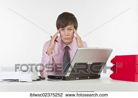 Stock Image - "Young man at his desk in the office with a telephone and a laptop computer, headache". Fotosearch