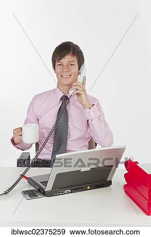 Stock Photo - "Young man at his desk with a laptop computer, making phone call, smiling". Fotosearch