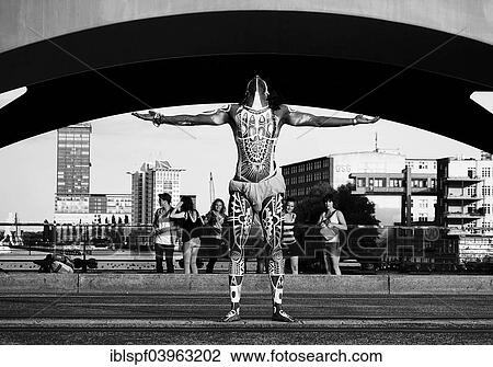 Stock Image - "Young man painted in Polynesian patterns standing with outstretched arms, Oberbaumbrucke bridge, Berlin, Germany, Europe". Fotosearch