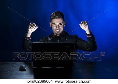 "Young man sitting in the dark in front of a laptop, with a joyful expression, exulting" View Large Photo Image Picture - "Young man sitting in the dark in front of a laptop, with a joyful expression, exulting". Fotosearch