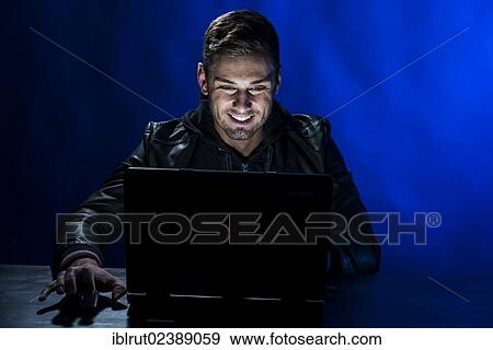 "Young man sitting in the dark in front of a laptop, with a joyful expression" View Large Photo Image Stock Photo - "Young man sitting in the dark in front of a laptop, with a joyful expression". Fotosearch