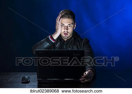 "Young man sitting in the dark in front of a laptop, with a shocked facial expression" View Large Photo Image Stock Photo - "Young man sitting in the dark in front of a laptop, with a shocked facial expression". Fotosearch