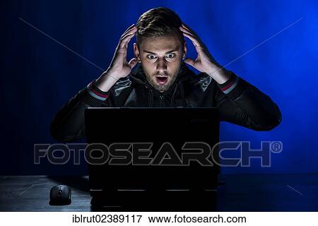 "Young man sitting in the dark in front of a laptop, with a shocked facial expression" View Large Photo Image Stock Photo - "Young man sitting in the dark in front of a laptop, with a shocked facial expression". Fotosearch