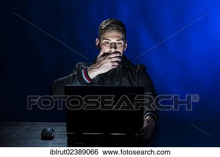 Stock Photograph - "Young man sitting in the dark in front of a laptop, with a shocked facial expression". Fotosearch