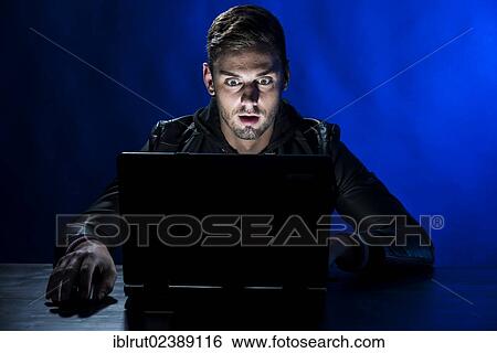 "Young man sitting in the dark in front of a laptop, with a shocked facial expression" View Large Photo Image Stock Photograph - "Young man sitting in the dark in front of a laptop, with a shocked facial expression". Fotosearch