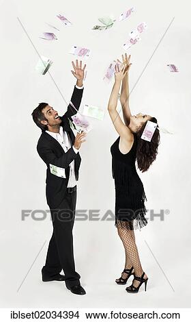 Picture - "Young man wearing a suit and a young woman in a black dress reaching joyfully for raining banknotes, symbolic image of success, profit". Fotosearch