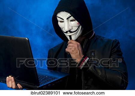 Stock Image - "Young man wearing an anonymous mask with a hood and a black leather jacket, holding a laptop, making a considering gesture". Fotosearch