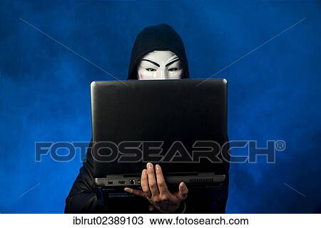 Stock Image - "Young man wearing an anonymous mask with a hood, holding a laptop". Fotosearch
