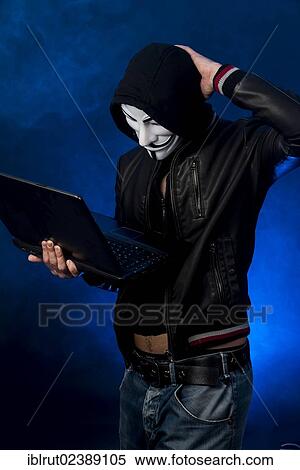 Stock Photography - "Young man wearing an anonymous mask with a hood and a black leather jacket, holding a laptop, scrathing his head and looking perplexed". Fotosearch