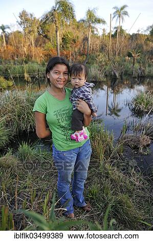 "Young mother holding a child in her arms in front of a pond in the community of Mbya-Guarani Indians, Campito, Caaguazu Department, Paraguay, South America" View Large Photo Image Stock Photo - "Young mother holding a child in her arms in front of a pond in the community of Mbya-Guarani Indians, Campito, Caaguazu Department, Paraguay, South America". Fotosearch