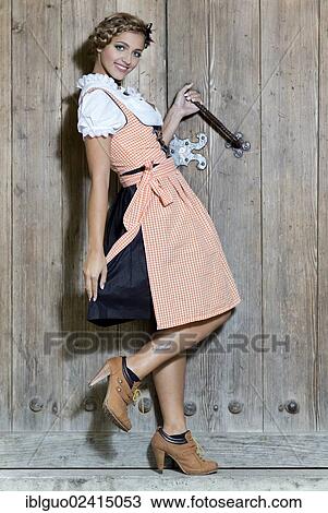 Stock Image - Young smiling woman wearing a dirndl dress posing against a wooden door, dirndl look. Fotosearch