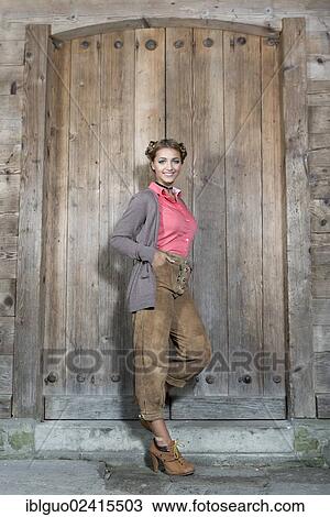 Stock Image - Young smiling woman wearing traditional leather pants posing in front of a wooden door, dirndl look. Fotosearch