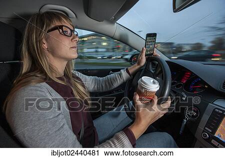 Stock Image - Young woman driving a car in the city centre while holding her cell phone and a cup of coffee at the same time. Fotosearch