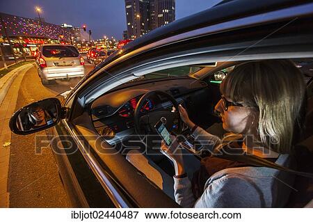 Young woman driving a car in the city centre in the evening while texting on her cell phone View Large Photo Image Stock Photo - Young woman driving a car in the city centre in the evening while texting on her cell phone. Fotosearch