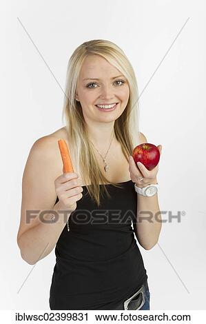 Stock Image - Young woman holding a carrot and an apple in her hands. Fotosearch