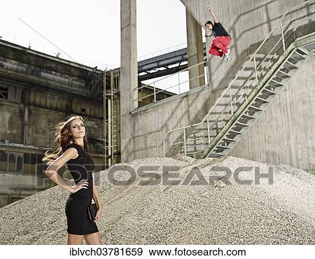 "Young woman in black dress standing in front of a cement plant, young man jumping from a railing, Kiefersfelden, Bavaria, Germany, Europe" View Large Photo Image Stock Photo - "Young woman in black dress standing in front of a cement plant, young man jumping from a railing, Kiefersfelden, Bavaria, Germany, Europe". Fotosearch