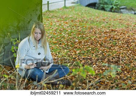 Stock Image - Young woman leaning against a tree in the park and looking at a tablet PC. Fotosearch