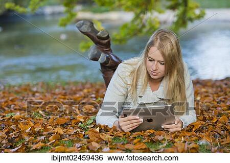 Stock Photograph - Young woman lying on her belly on the ground in a park and looking at a tablet PC. Fotosearch