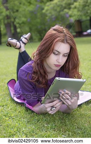Young woman lying on the grass in a park and reading on a tablet pc View Large Photo Image Stock Photography - Young woman lying on the grass in a park and reading on a tablet pc. Fotosearch