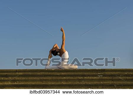 Picture - "Young woman practising Hatha yoga, outdoors, showing the pose Kapotasana, Pigeon pose". Fotosearch