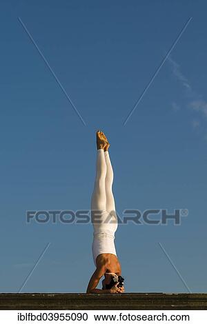 Stock Image - "Young woman practising Hatha yoga, outdoors, showing the pose Sirshasana, head stand". Fotosearch