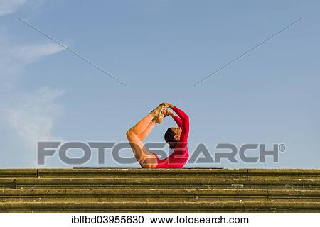 Stock Image - "Young woman practising Hatha yoga, outdoors, showing the pose Dhanurasana, Bow pose". Fotosearch