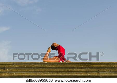 Stock Image - "Young woman practising Hatha yoga, outdoors, showing the pose Kapotasana, Pigeon pose". Fotosearch
