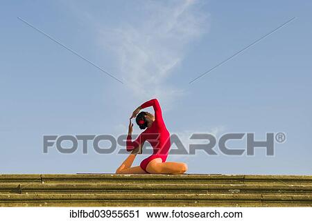 Stock Image - "Young woman practising Hatha yoga, outdoors, showing the pose Kapotasana, Pigeon pose". Fotosearch