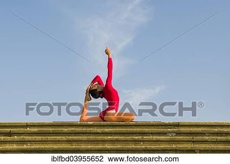 Stock Image - "Young woman practising Hatha yoga, outdoors, showing the pose Kapotasana, Pigeon pose". Fotosearch