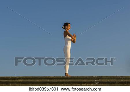Stock Image - "Young woman practising Hatha yoga, outdoors, showing the pose Tadasana, Pranamasana, Prayer pose". Fotosearch