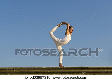 Stock Image - "Young woman practising Hatha yoga, outdoors, showing the pose Natarajasana, Lord Shiva's pose". Fotosearch