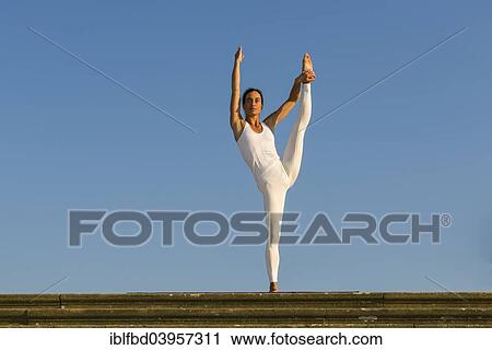 Stock Image - "Young woman practising Hatha yoga, outdoors, showing the pose Anjaneyasana, Raised hand to foot pose". Fotosearch