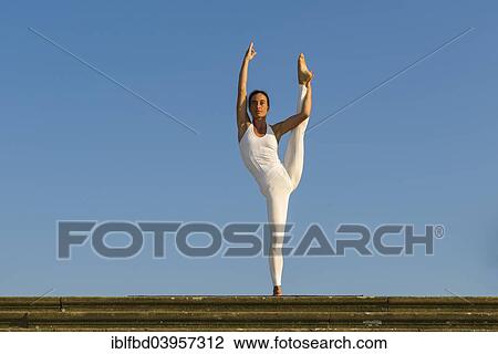 Stock Image - "Young woman practising Hatha yoga, outdoors, showing the pose Anjaneyasana, Raised hand to foot pose". Fotosearch