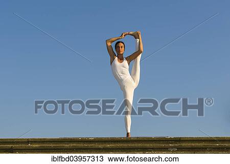 Stock Image - "Young woman practising Hatha yoga, outdoors, showing the pose Utthita hasta pandangusthasana, Raised hand to big toe pose". Fotosearch