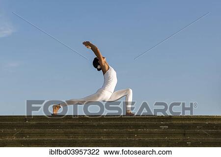 Stock Image - "Young woman practising Hatha yoga, outdoors, showing the pose Anjaneyasana, Chandrasana, Half moon pose". Fotosearch