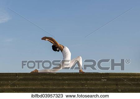 Stock Image - "Young woman practising Hatha yoga, outdoors, showing the pose Anjaneyasana, Chandrasana, Half moon pose". Fotosearch