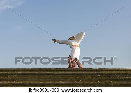 Stock Image - "Young woman practising Hatha yoga, outdoors, showing the pose Sirshasana, Head stand". Fotosearch