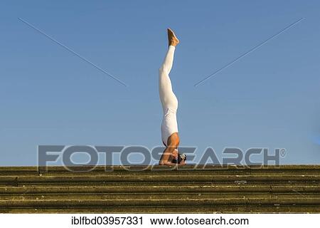 Stock Image - "Young woman practising Hatha yoga, outdoors, showing the pose Sirshasana, Head stand". Fotosearch