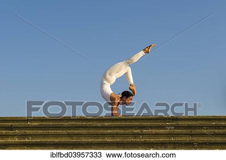 Stock Image - "Young woman practising Hatha yoga, outdoors, showing the pose Vrischikasana, Scorpion pose". Fotosearch