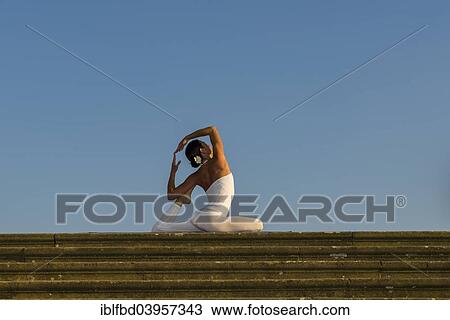 Stock Image - "Young woman practising Hatha yoga, outdoors, showing the pose Kapotasana, Pigeon pose". Fotosearch