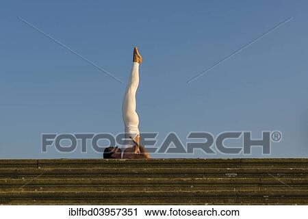 Stock Image - "Young woman practising Hatha yoga, outdoors, showing the pose Sarvangasana, Shoulderstand". Fotosearch