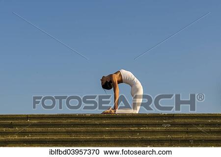 Stock Image - "Young woman practising Hatha yoga, outdoors, showing the pose Ushtrasana, Camel pose". Fotosearch