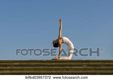 Stock Image - "Young woman practising Hatha yoga, outdoors, showing the pose Ushtrasana, Camel pose". Fotosearch