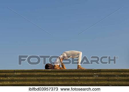Stock Image - "Young woman practising Hatha yoga, outdoors, showing the pose variation of Halasana, Supta konasana, variation of Plough". Fotosearch