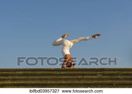 Stock Photo - "Young woman practising Hatha yoga, outdoors, showing the pose Sirshasana, Head stand". Fotosearch