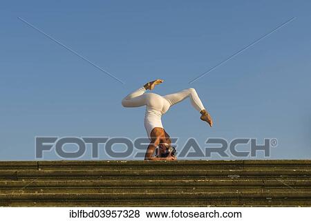 Stock Photo - "Young woman practising Hatha yoga, outdoors, showing the pose Sirshasana, Head stand". Fotosearch